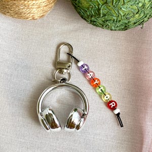 May include: A silver headphone keychain with a lobster clasp. The keychain has a beaded charm with the name "Taylor" spelled out in colorful letter beads. The headphones are detailed with ear cups.