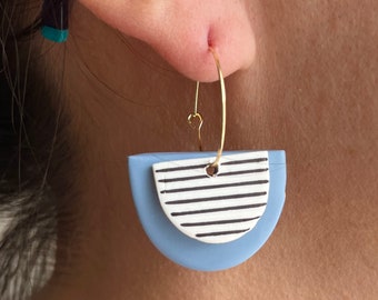 May include: A pair of earrings with a gold-colored hook. The earrings have a light blue, semi-circular base, topped with a white semi-circle featuring black horizontal stripes. The earrings are worn on the ear.