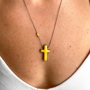 Op de afbeelding: Een ketting met een hanger in de vorm van een geel keramisch kruis aan een dun zwart koord. De ketting wordt door een persoon gedragen.