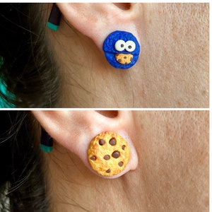 May include: A pair of earrings featuring a blue cookie monster and a chocolate chip cookie. The cookie monster earring has white eyes and a small cookie in its mouth. The chocolate chip cookie earring is brown with white chocolate chips.