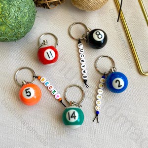 May include: Keychains featuring miniature billiard balls in various colors, each with a different number. Some keychains have personalized beaded name tags. The keychains are attached to silver-colored key rings.