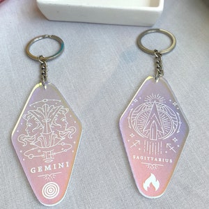 May include: Two iridescent keychains with silver chains. The keychains are shaped like motel signs. One keychain has the zodiac sign Gemini with the text "GEMINI" and the other keychain has the zodiac sign Sagittarius with the text "SAGITTARIUS".