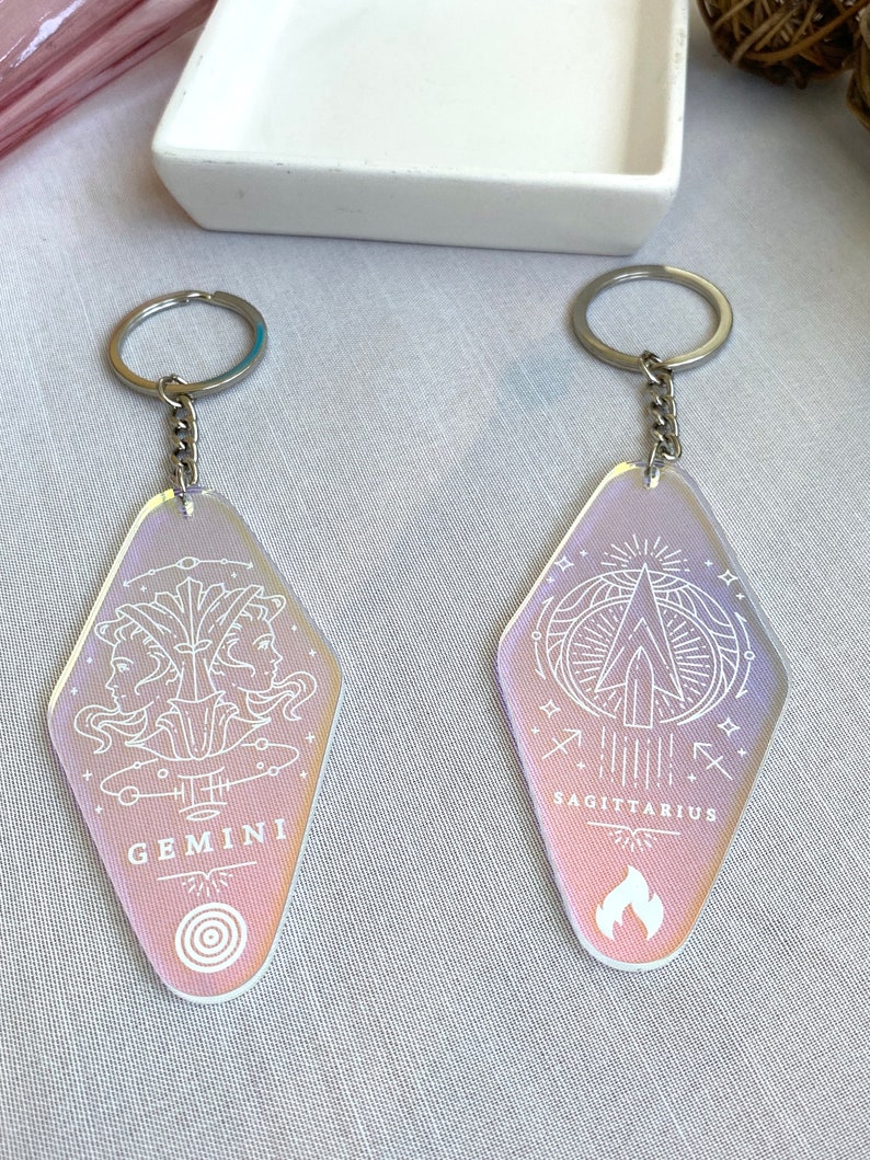 May include: Two iridescent keychains with silver chains. The keychains are shaped like motel signs. One keychain has the zodiac sign Gemini with the text "GEMINI" and the other keychain has the zodiac sign Sagittarius with the text "SAGITTARIUS".