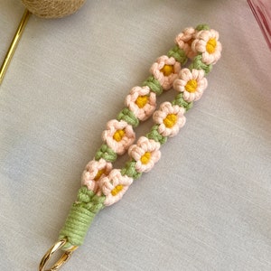 May include: A pink and green crocheted keychain with a gold metal ring. The keychain is decorated with small pink flowers with yellow centers.