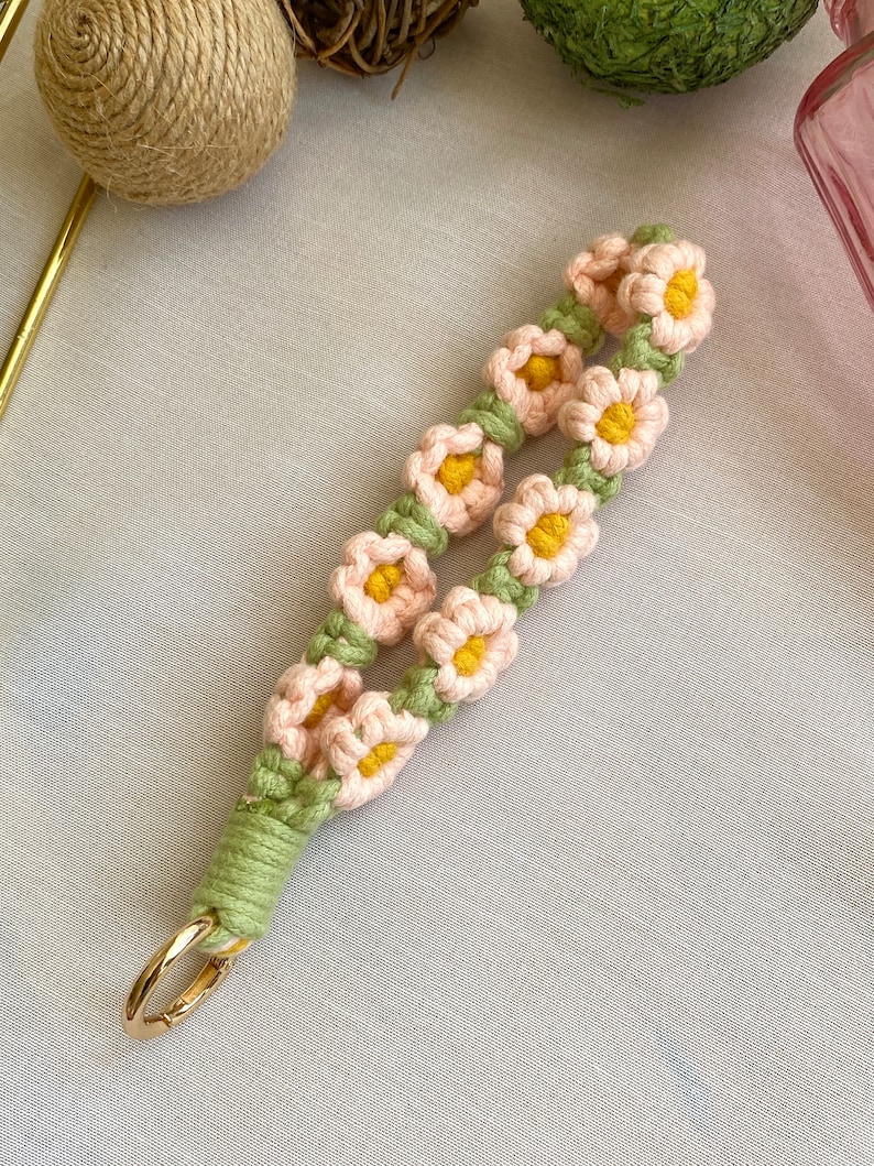 May include: A pink and green crocheted keychain with a gold metal ring. The keychain is decorated with small pink flowers with yellow centers.