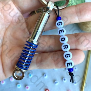 May include: A silver car shock absorber keychain with a blue spring. The keychain also has a black string with white beads that spell out the name "GEORGE".