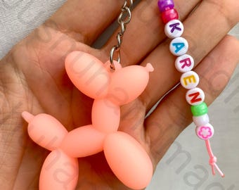 Könnte beinhalten: Ein rosa Ballonhund-Schlüsselanhänger mit einem silbernen Schlüsselring. Der Schlüsselanhänger enthält eine Perlenkette mit dem Namen "KAREN" in weißen Buchstabenperlen, akzentuiert mit bunten Perlen und einem Blumenanhänger.