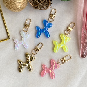 May include: Five colorful balloon dog keychains. The keychains are made of iridescent plastic and come in blue, yellow, pink, white, and gold. Each keychain features a gold metal ring and a lobster clasp.