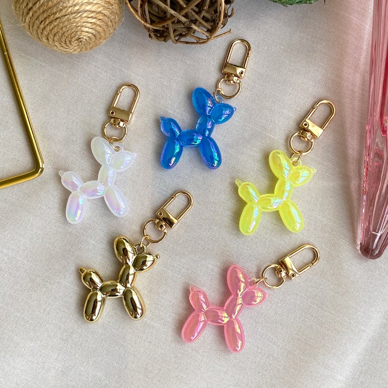 May include: Five colorful balloon dog keychains. The keychains are made of iridescent plastic and come in blue, yellow, pink, white, and gold. Each keychain features a gold metal ring and a lobster clasp.