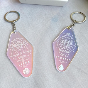 May include: Two iridescent keychains with silver rings. The keychain on the left features a Libra zodiac symbol with the word "LIBRA" printed below. The keychain on the right features a Scorpio zodiac symbol with the word "SCORPIO" printed below.