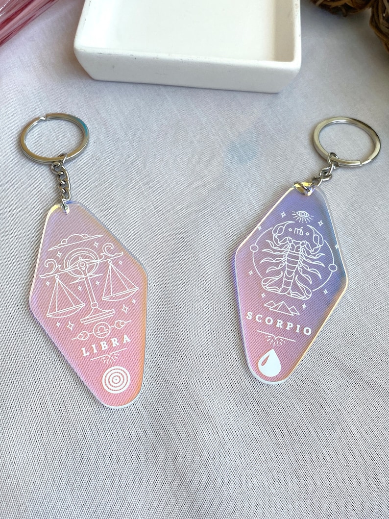 May include: Two iridescent keychains with silver rings. The keychain on the left features a Libra zodiac symbol with the word "LIBRA" printed below. The keychain on the right features a Scorpio zodiac symbol with the word "SCORPIO" printed below.
