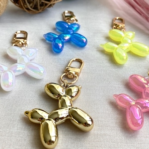 May include: Five balloon dog keychains in different colors. The keychains are made of a clear plastic material with a shiny finish. The colors are iridescent white, blue, yellow, pink, and gold.