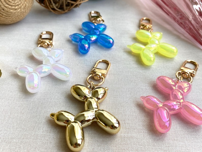 May include: Five balloon dog keychains in different colors. The keychains are made of a clear plastic material with a shiny finish. The colors are iridescent white, blue, yellow, pink, and gold.