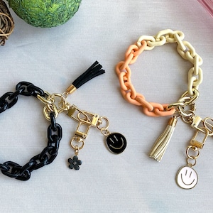 May include: Two keychains with black and white smiley face charms. One keychain has a black chain with a black tassel and the other has a multi-colored chain with a cream tassel.