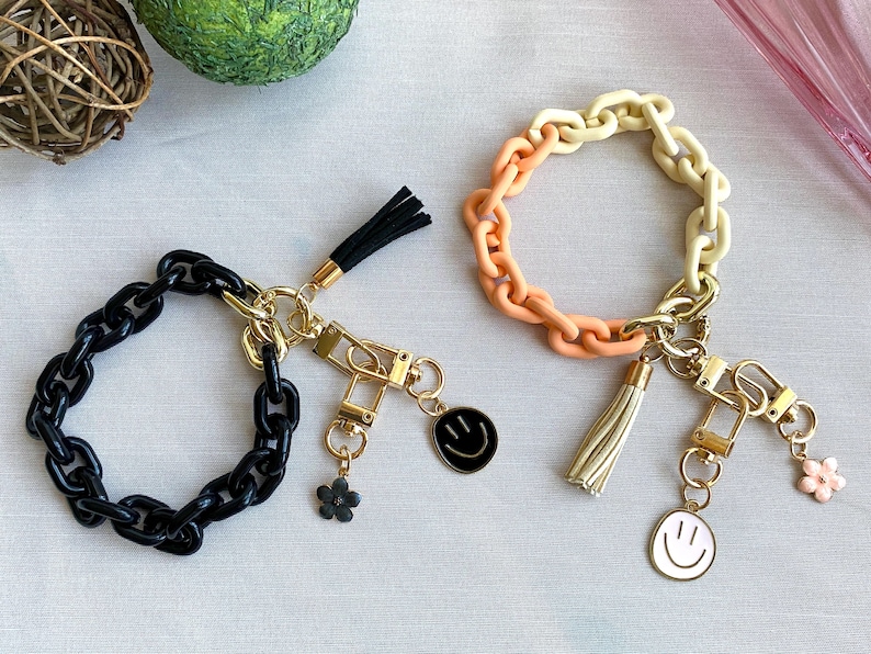 May include: Two keychains with black and white smiley face charms. One keychain has a black chain with a black tassel and the other has a multi-colored chain with a cream tassel.