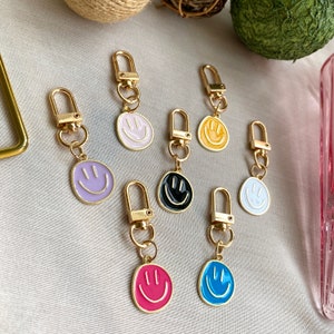 May include: A set of seven gold keychains with different colored enamel smiley faces. The keychains are all attached to gold rings.