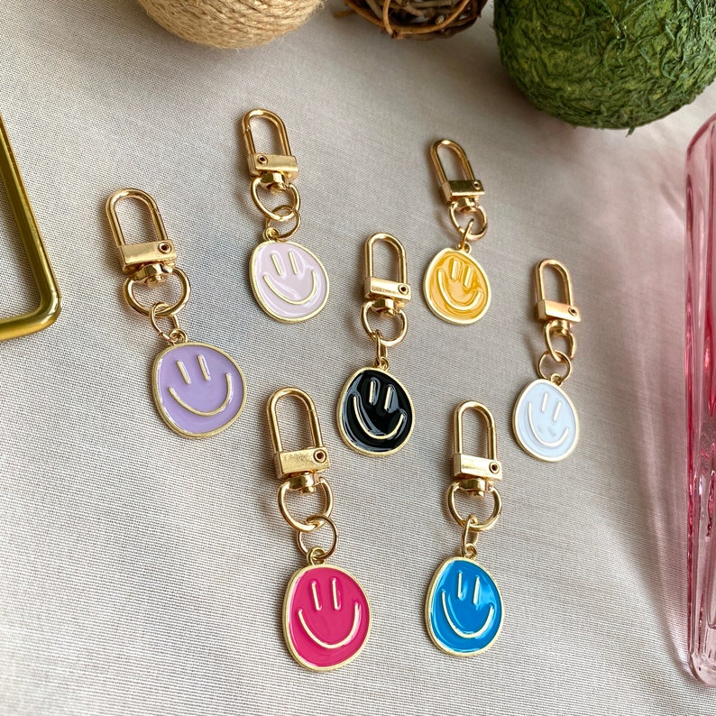 May include: A set of seven gold keychains with different colored enamel smiley faces. The keychains are all attached to gold rings.