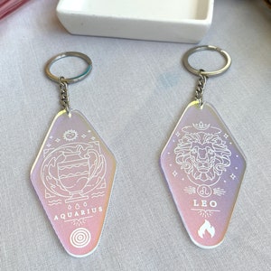May include: Two iridescent keychains with silver key rings. The keychains are shaped like a motel sign and feature the zodiac signs Aquarius and Leo with their corresponding symbols. The Aquarius keychain has a water pitcher and waves, while the Leo keychain has a lion's head and flames.