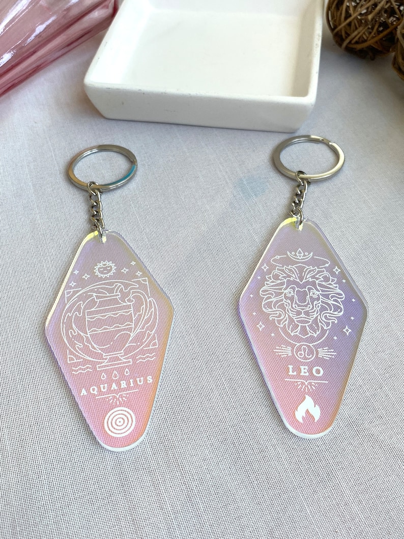 May include: Two iridescent keychains with silver key rings. The keychains are shaped like a motel sign and feature the zodiac signs Aquarius and Leo with their corresponding symbols. The Aquarius keychain has a water pitcher and waves, while the Leo keychain has a lion's head and flames.
