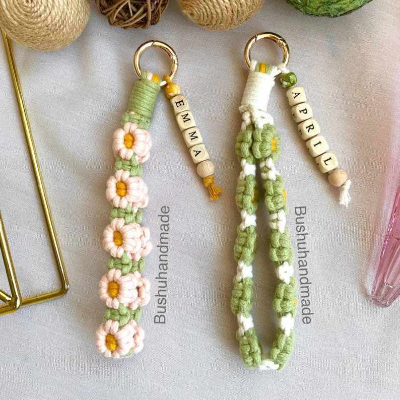 May include: Two green and white crocheted keychains with wooden beads spelling out the names "Emma" and "April". The keychains feature a daisy design and are labeled "Bushuhhandmade".