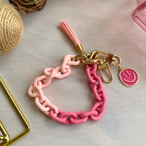 May include: A pink and light pink chain keychain with a gold charm and a pink smiley face charm. The keychain has a pink tassel.
