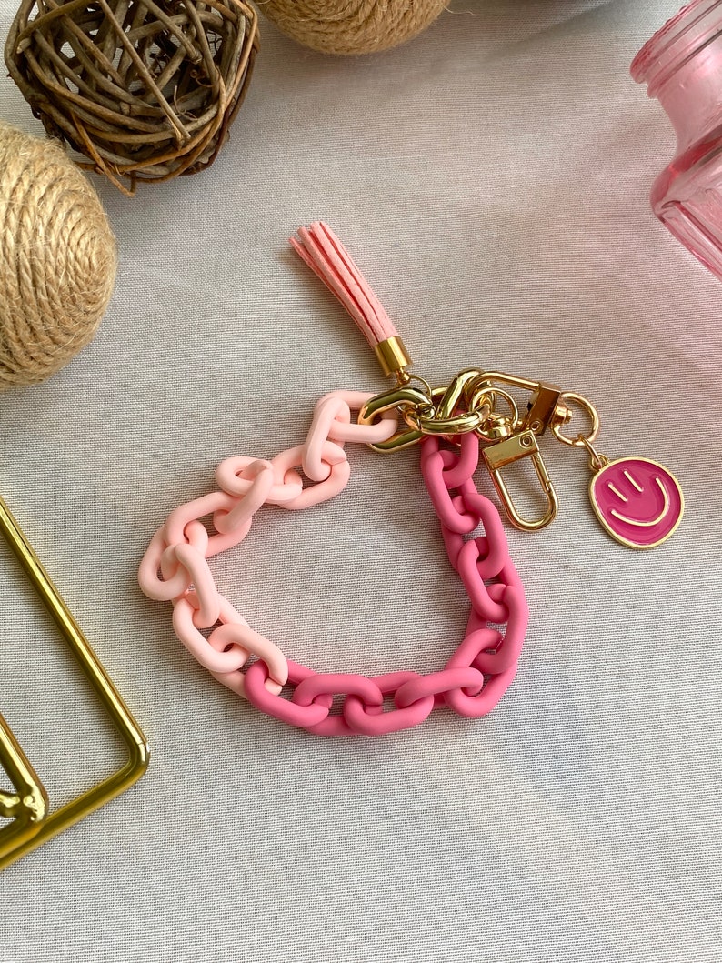 May include: A pink and light pink chain keychain with a gold charm and a pink smiley face charm. The keychain has a pink tassel.
