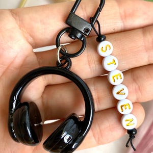 Puede incluir: Llavero de auriculares negros con el nombre "STEVEN" en letras doradas sobre cuentas blancas. El llavero tiene un mosquet&oacute;n negro y un peque&ntilde;o cord&oacute;n negro.