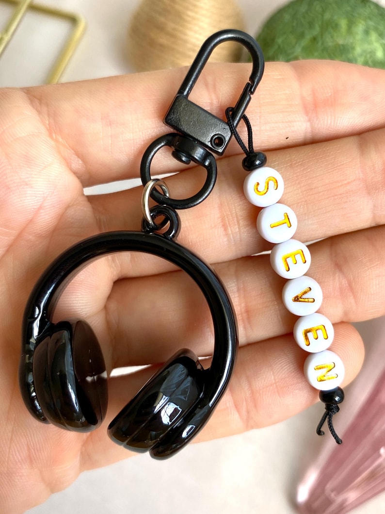 Puede incluir: Llavero de auriculares negros con el nombre "STEVEN" en letras doradas sobre cuentas blancas. El llavero tiene un mosquet&oacute;n negro y un peque&ntilde;o cord&oacute;n negro.
