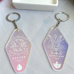 May include: Two iridescent keychains with silver key rings. The keychains are shaped like a diamond and feature white line drawings of the zodiac signs Pisces and Capricorn. The Pisces keychain has a fish and the Capricorn keychain has a goat.
