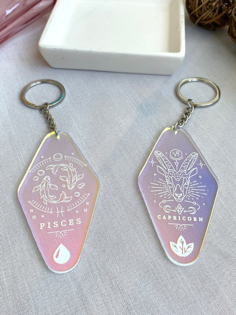 May include: Two iridescent keychains with silver key rings. The keychains are shaped like a diamond and feature white line drawings of the zodiac signs Pisces and Capricorn. The Pisces keychain has a fish and the Capricorn keychain has a goat.