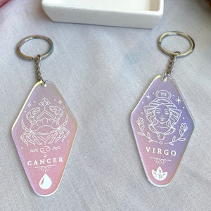 May include: Two iridescent keychains with silver key rings. The keychains are shaped like a vintage motel sign. One keychain features the zodiac sign Cancer with a crab and the word "Cancer". The other keychain features the zodiac sign Virgo with a woman's face and the word "Virgo".
