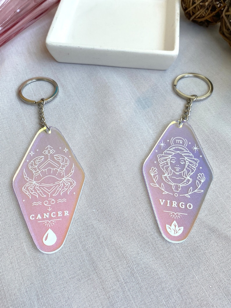 May include: Two iridescent keychains with silver key rings. The keychains are shaped like a vintage motel sign. One keychain features the zodiac sign Cancer with a crab and the word "Cancer". The other keychain features the zodiac sign Virgo with a woman's face and the word "Virgo".