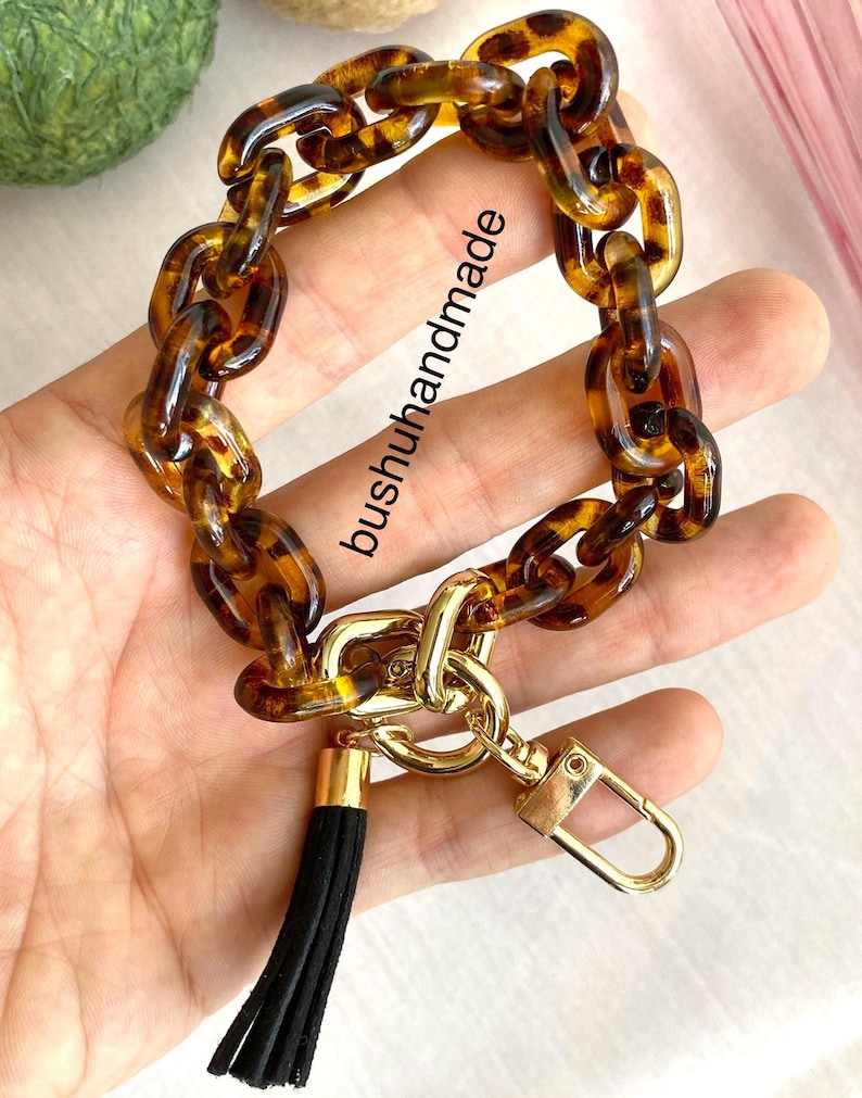 May include: A brown and gold chain keychain with a black tassel. The chain is made of large, chunky links with a tortoise shell pattern. The keychain has a gold clasp and a gold ring.