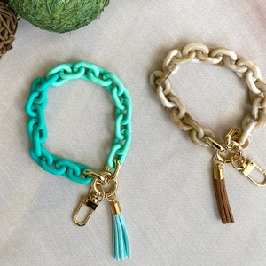 May include: Two colorful chain bracelets with gold keychains and tassels. One bracelet is a gradient of teal and green, the other is a light brown color.