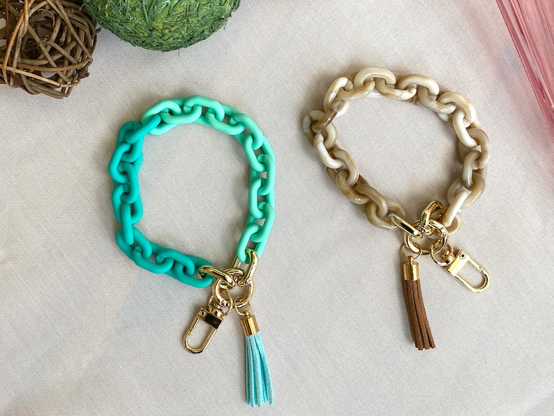 May include: Two colorful chain bracelets with gold keychains and tassels. One bracelet is a gradient of teal and green, the other is a light brown color.