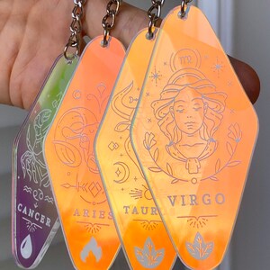 May include: Four iridescent orange keychains with zodiac signs: Cancer, Aries, Taurus, and Virgo. Each keychain features a different zodiac symbol and the name of the sign.