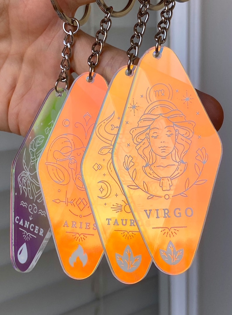May include: Four iridescent orange keychains with zodiac signs: Cancer, Aries, Taurus, and Virgo. Each keychain features a different zodiac symbol and the name of the sign.
