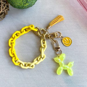 May include: A yellow and light yellow plastic chain keychain with a gold metal charm of a smiley face and a green balloon animal shaped like a dog.