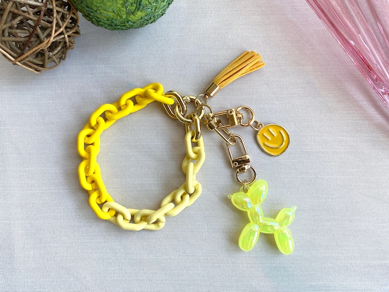 May include: A yellow and light yellow plastic chain keychain with a gold metal charm of a smiley face and a green balloon animal shaped like a dog.