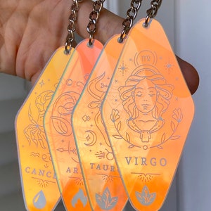 May include: Four iridescent keychains with zodiac signs: Cancer, Aries, Taurus, and Virgo. Each keychain features a different zodiac symbol and a corresponding element: water, fire, earth, and air.