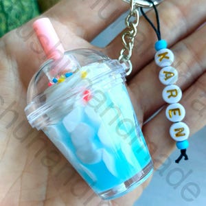 May include: A miniature blue and white drink keychain with a clear lid and pink straw. The keychain includes a silver chain and a beaded charm spelling "KAREN". The drink has colourful sprinkles.