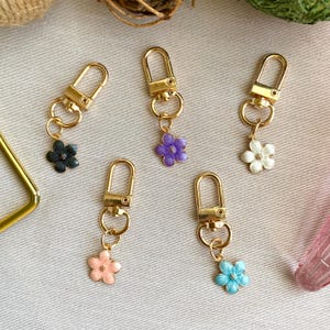 May include: Five gold keychains with different colored flower charms. The charms are purple, white, black, pink, and blue.