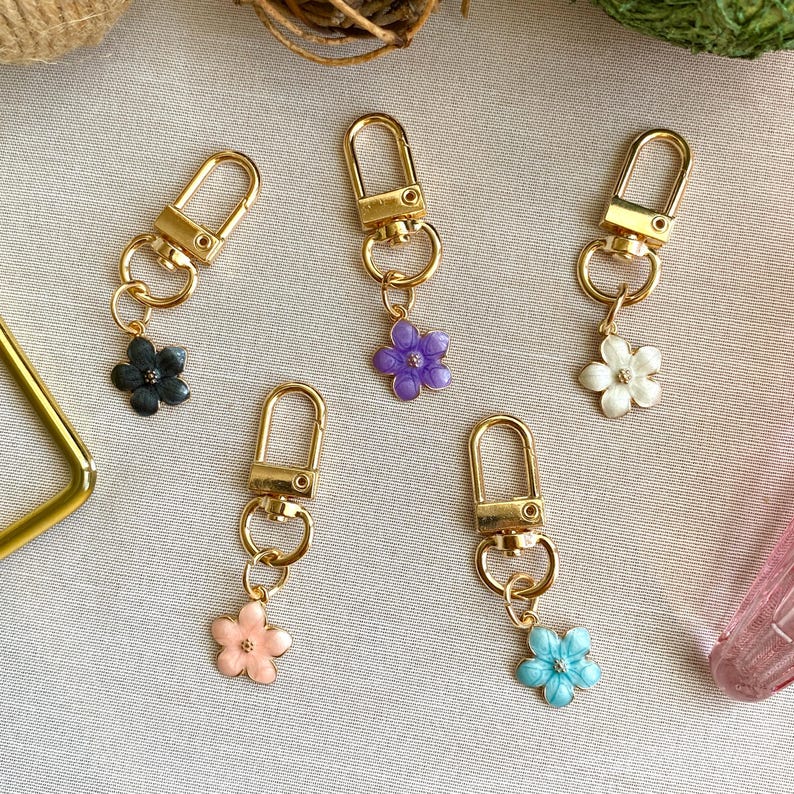May include: Five gold keychains with different colored flower charms. The charms are purple, white, black, pink, and blue.