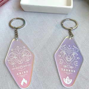 May include: Two iridescent keychains with silver key rings. The keychains are shaped like a retro motel sign. The left keychain features a white line drawing of an Aries zodiac symbol with the word "ARIES" below. The right keychain features a white line drawing of a Taurus zodiac symbol with the word "TAURUS" below.