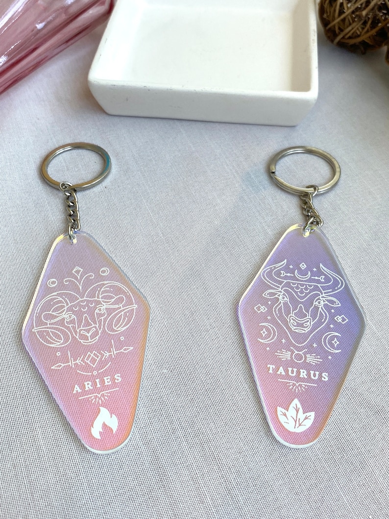 May include: Two iridescent keychains with silver key rings. The keychains are shaped like a retro motel sign. The left keychain features a white line drawing of an Aries zodiac symbol with the word "ARIES" below. The right keychain features a white line drawing of a Taurus zodiac symbol with the word "TAURUS" below.