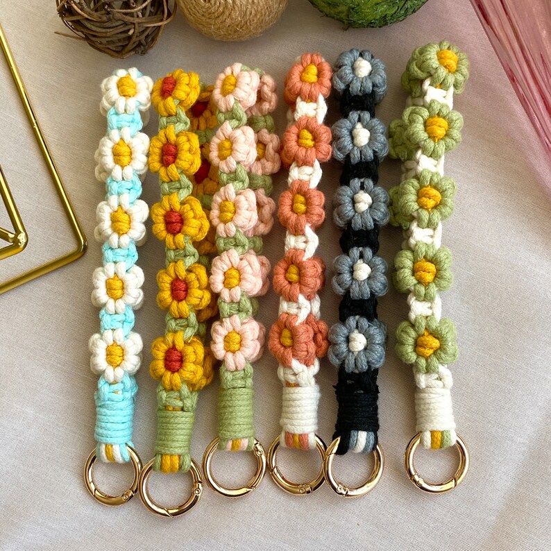 May include: Six colorful crocheted keychains with flower designs. Each keychain has a gold metal ring attached. The keychains are in the colors blue, yellow, pink, orange, gray, and green.