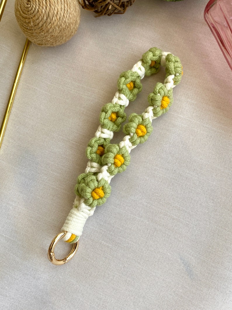 May include: A green and white crocheted keychain with a gold metal ring. The keychain is decorated with small yellow flowers.