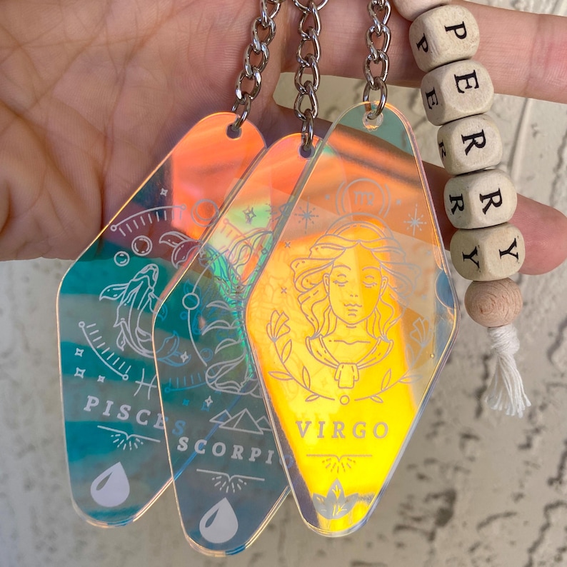 May include: Three iridescent keychains with zodiac signs: Pisces, Scorpio, and Virgo. Each keychain is shaped like a motel sign and has a silver chain.
