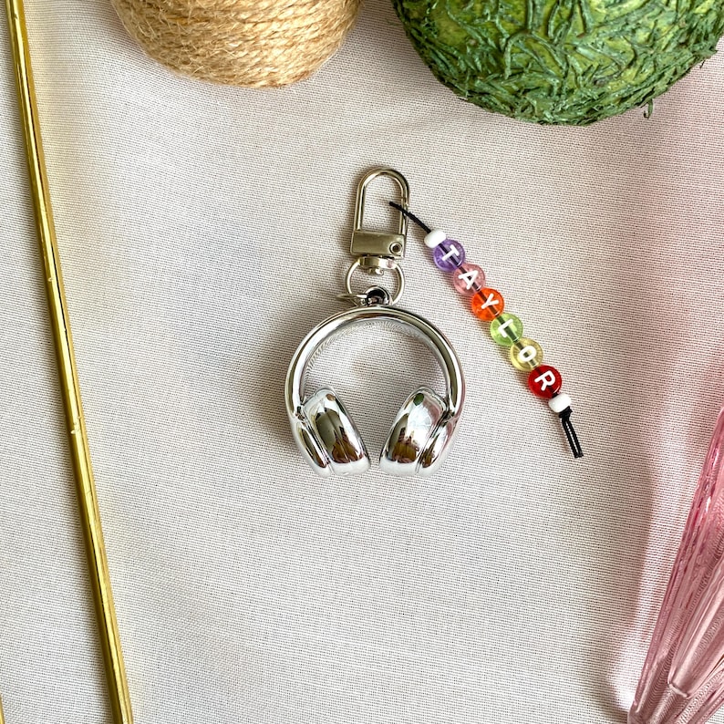 May include: A silver headphone keychain with a lobster clasp. The keychain has a beaded charm with the name "Taylor" spelled out in colorful letter beads. The headphones are detailed with ear cups.