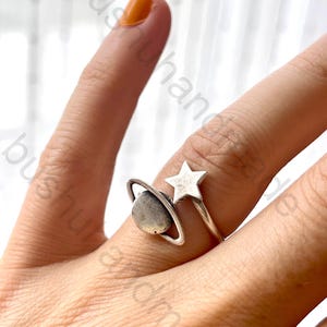 May include: A silver ring featuring a planet and star design. The ring has a small, round planet with a silver band around it, and a five-pointed star. The ring is worn on a finger with orange nail polish.